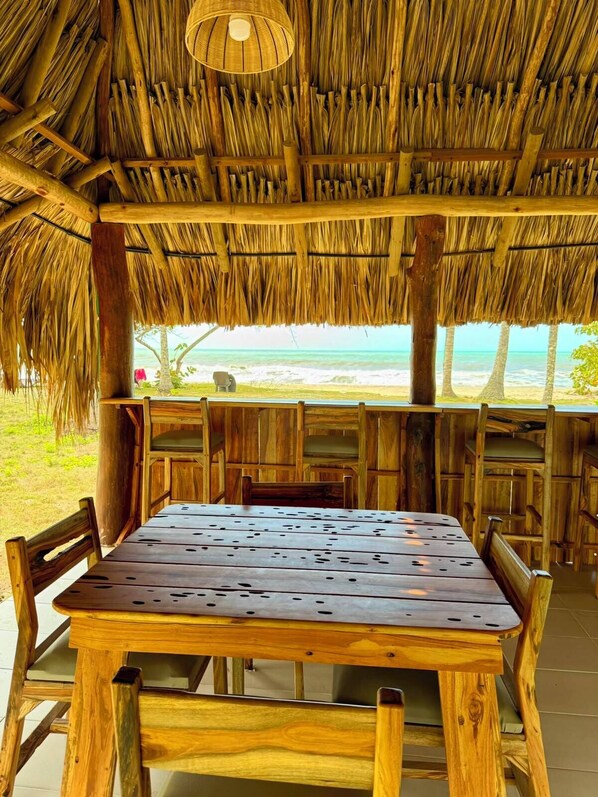 Mesa y sillas de madera bajo un techo de paja, con vistas a la playa.