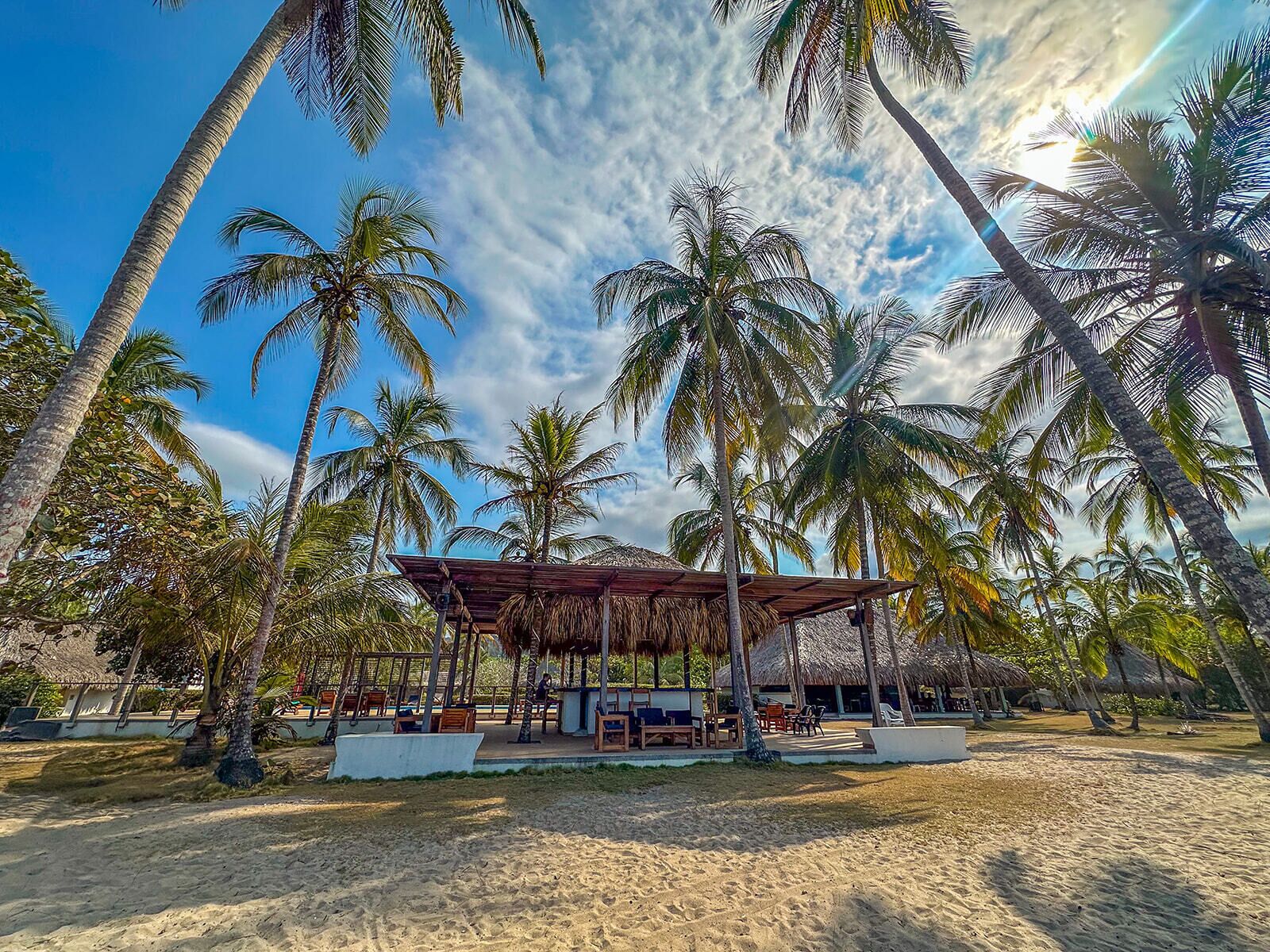 A beachside resort with thatched-roof structures, palm trees, and sandy ground.