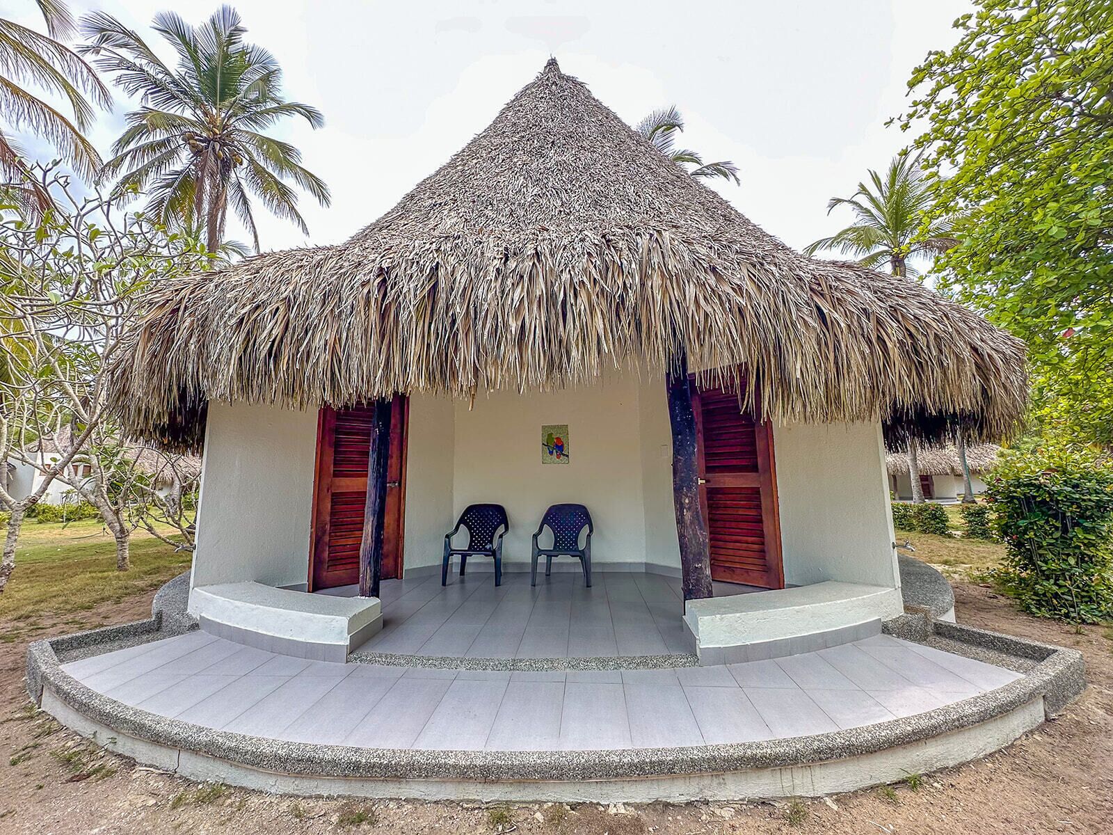 A thatched-roof hut with two wooden doors and two plastic chairs in front.