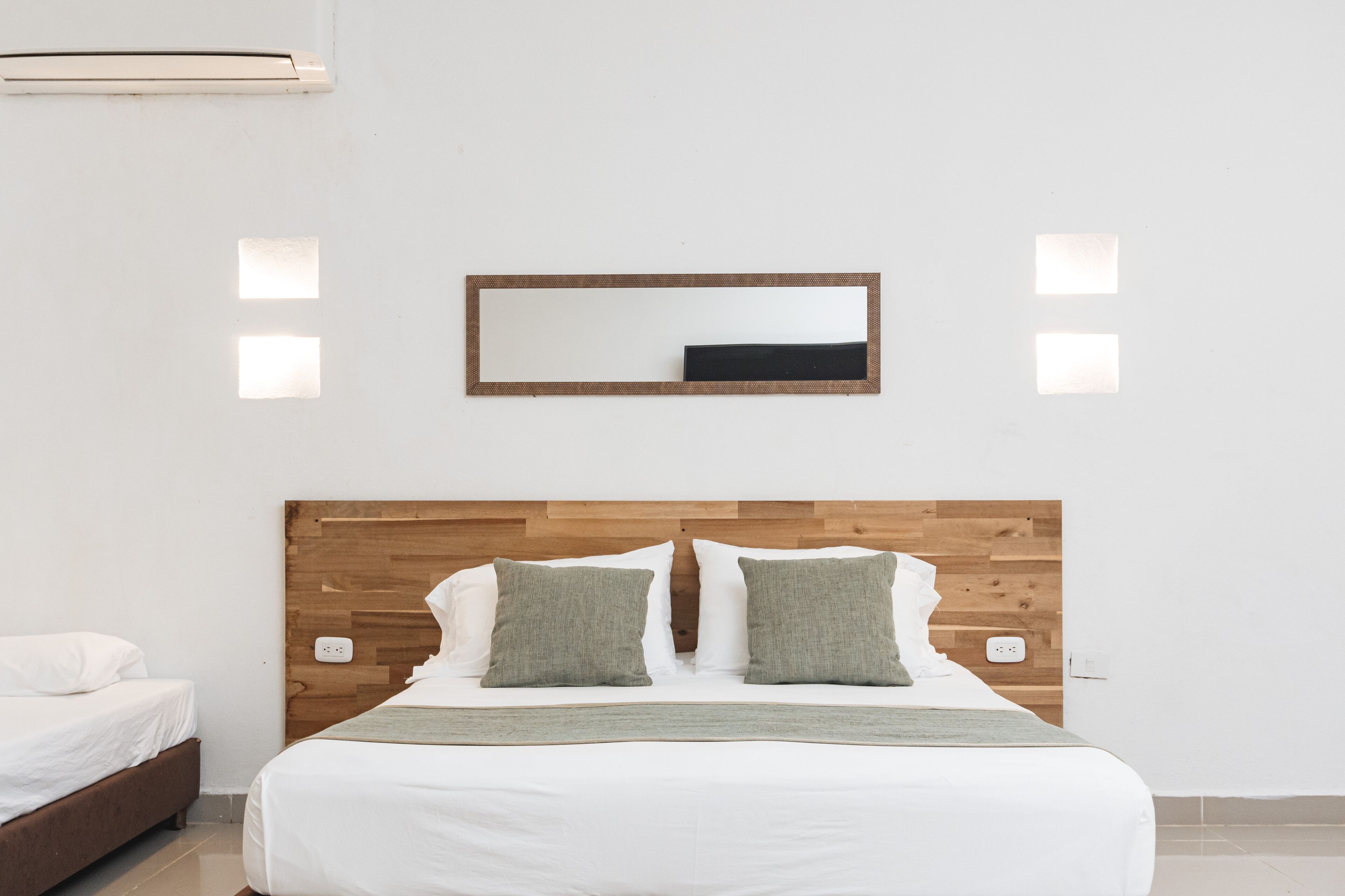 A bedroom with a wooden headboard, two pillows, a bedside table, and a mirror on the wall.
