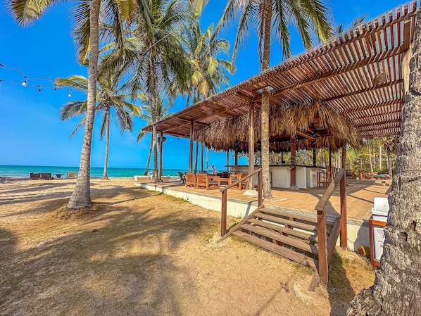 Un resort frente a la playa con una zona de estar cubierta, palmeras y vistas al océano.