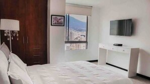 Een slaapkamer met een bed, bureau, televisie en raam met uitzicht op de stad.