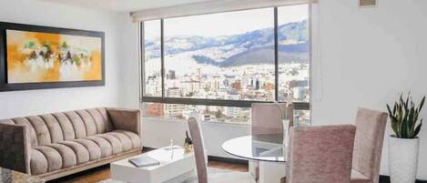 A modern living room with a sofa, a coffee table, and a dining area with chairs. A large window offers a city view.
