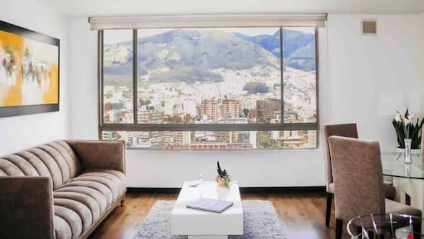 Een moderne woonkamer met een groot raam dat uitzicht over de stad biedt, een bank, een bijzettafeltje en twee fauteuils.