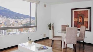 Een moderne woonkamer met een groot raam, een eettafel met stoelen, een salontafel en een kunstwerk in een lijst aan de muur.