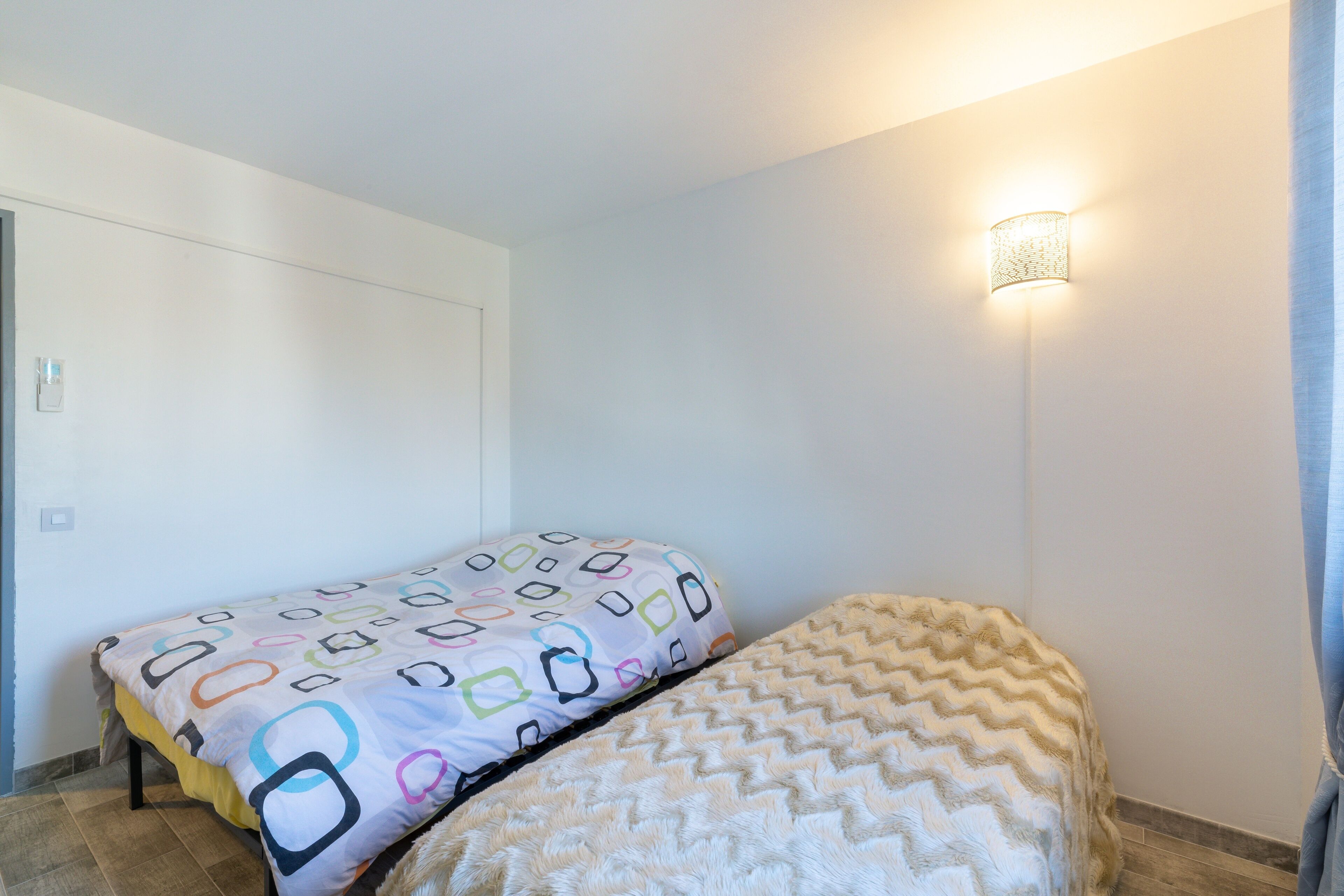 Una stanza con due letti, uno con una coperta dai colori vivaci e motivi geometrici e l'altro con una coperta a zig-zag. Sulla parete destra è presente un punto luce fissato al muro.