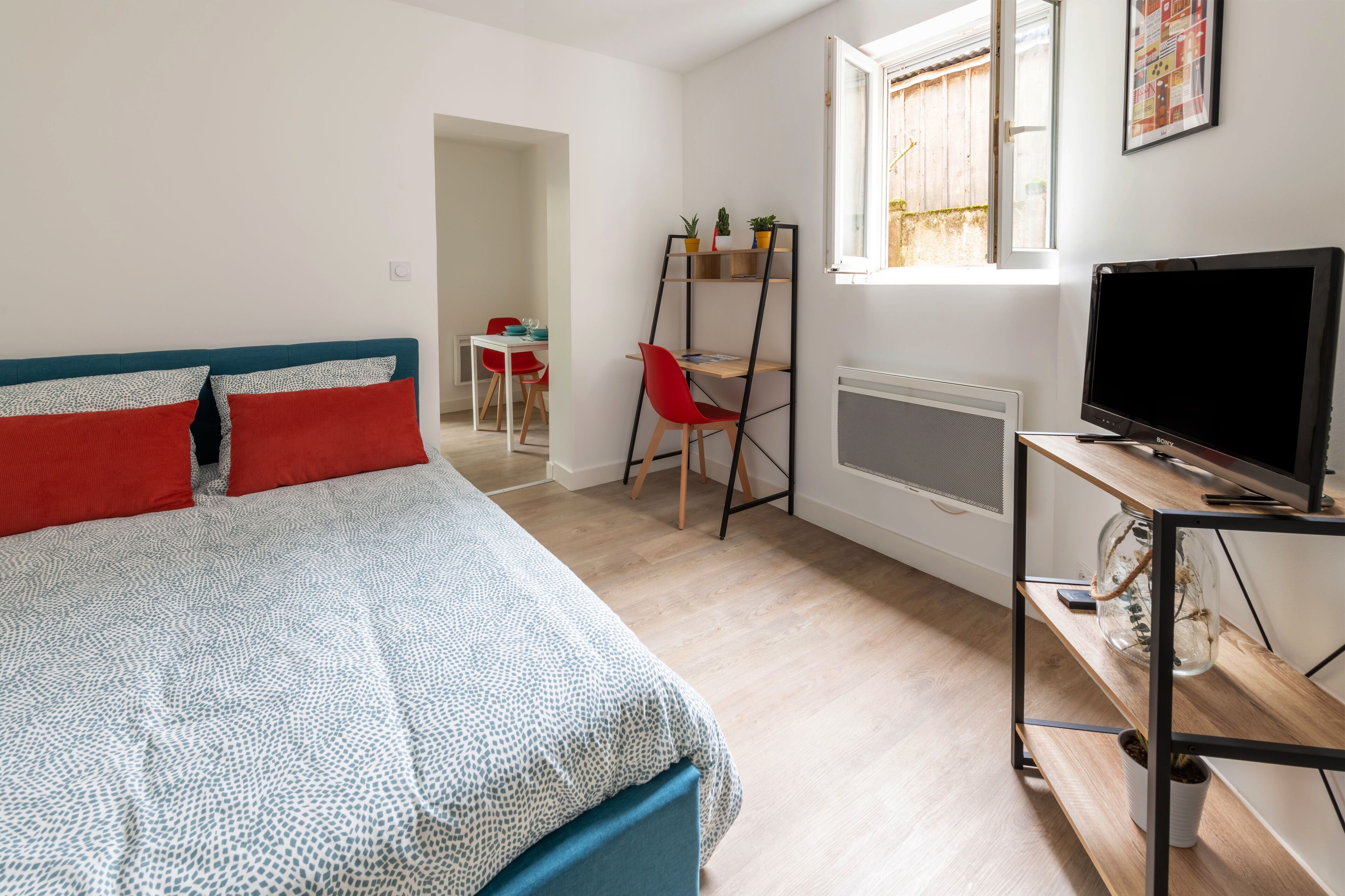 A bedroom with a bed, a TV, a shelf with plants, and a window.