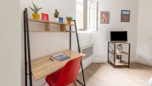 Una habitación pequeña con un escritorio de madera, una silla roja, un televisor sobre un mueble, y estanterías con plantas y libros.