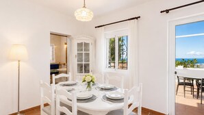 Uma área de refeições com uma mesa branca posta para quatro pessoas, um candelabro e vista para o mar pela janela.