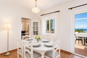 A dining area with a white table set for four, a chandelier, and a view of the sea through the window.