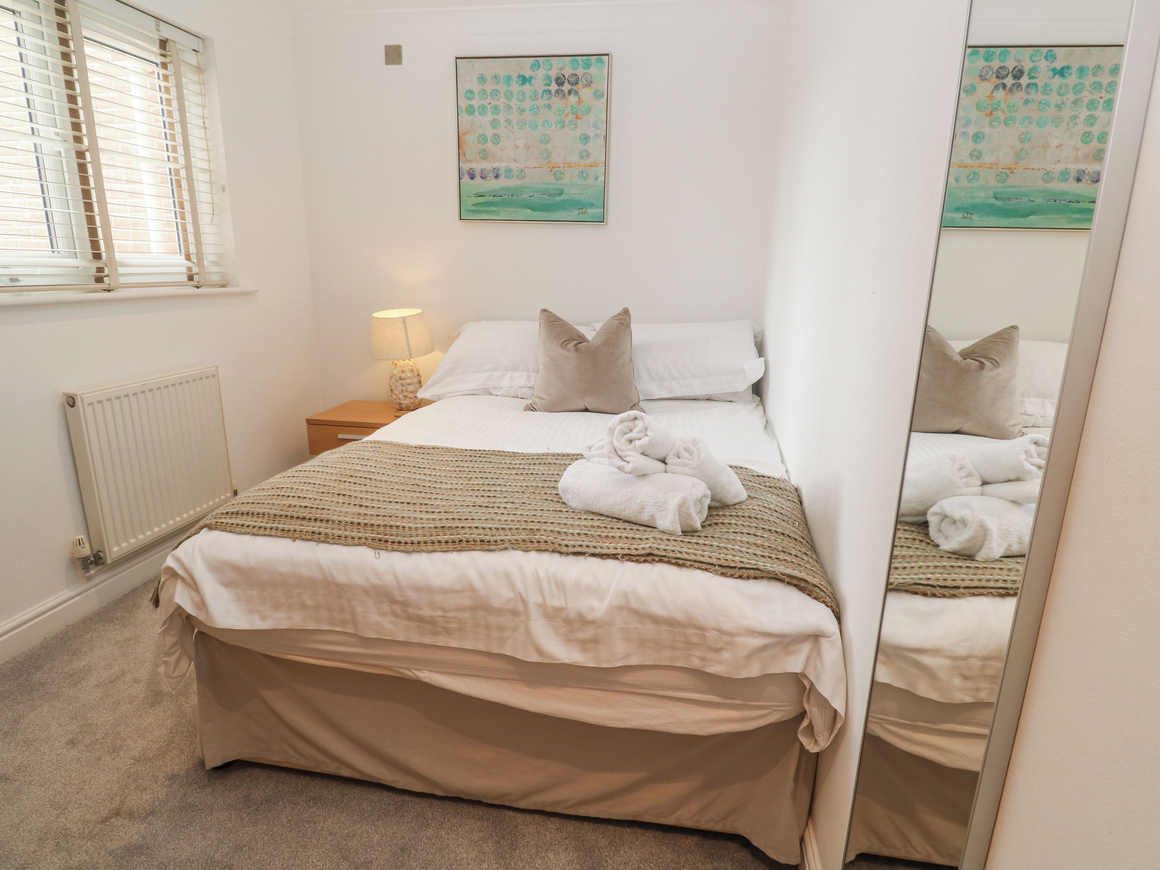 Un dormitorio ordenado con una cama, una mesita de noche con una lámpara, un radiador y dos cuadros colgados en la pared.