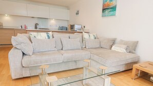 Un salon moderne avec un canapé d’angle aux couleurs claires, une table basse en verre et une table d’appoint en bois.