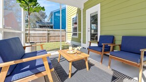 Eine überdachte Terrasse mit Holzmöbeln, einem Tisch mit Getränken und Blick auf ein blaues Haus und einen Garten.