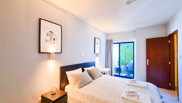 Une chambre à coucher bien rangée, avec un grand lit, une table de chevet, un tableau encadré et une vue sur l’extérieur par une fenêtre.