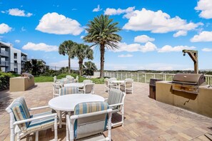 Un patio exterior con mesas redondas y sillas a rayas, una parrilla y palmeras.