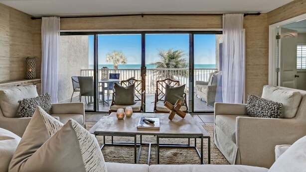 Una sala de estar moderna con un gran ventanal corredera que da a un balcón con vista al mar. Hay dos sillones, un sofá y una mesa de centro con objetos decorativos.