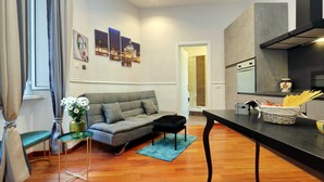 A modern living room with a grey sofa, a small table with a vase of flowers, a kitchen area with a stove and a basket of pasta. - 4bnb - Otranto Vatican Apartment (Rome)