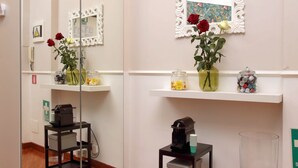 A hotel lobby with a mirror, a framed painting, and vases with flowers. There are shelves with various items and a small table with a coffee machine. - 4bnb - Otranto Vatican Apartment (Rome)