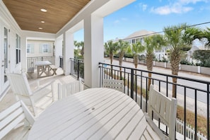 A balcony with a table and chairs, a wooden ceiling, and a view of palm trees and a building in the background. - 30A Paradise! Pool, Tiki Bar & 6-Seat Golf Cart 5-Bed Book Now for Fall! (Santa Rosa Beach)