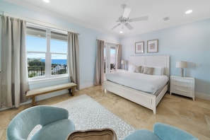 A bedroom with a large bed, a ceiling fan, a bench, two chairs, and a view of the ocean through a window. - 30A Paradise! Pool, Tiki Bar & 6-Seat Golf Cart 5-Bed Book Now for Fall! (Santa Rosa Beach)