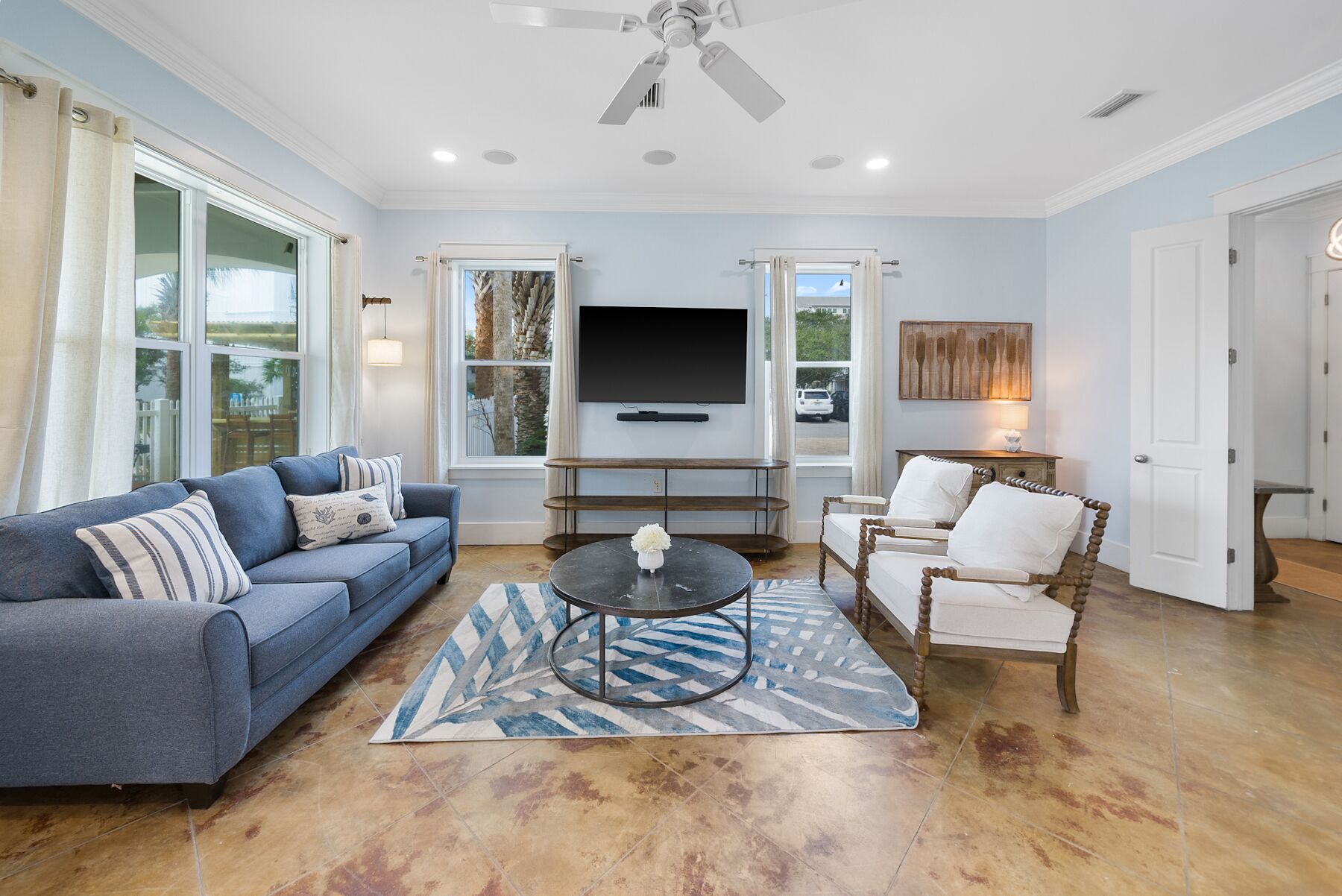 A spacious living room with a blue sofa, a coffee table, and a wooden chair. A flat-screen TV is mounted on the wall, and there is a window with curtains.