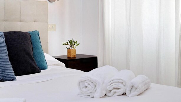 Een net opgemaakt bed met witte lakens, een klein nachtkastje met een plantje en een wandlamp.