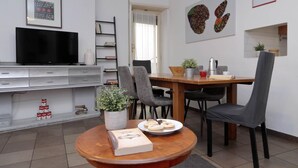Een moderne woonkamer met een flatscreen televisie, een eettafel met stoelen, een salontafel met boeken en een bord koekjes, en een boekenkast.