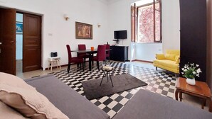 Un soggiorno con pavimento a scacchi, un divano, una poltrona gialla, un tavolo da pranzo con sedie, una TV e un quadro appeso al muro.