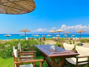 A beachside seating area with wooden tables and chairs, surrounded by thatched umbrellas, and overlooking the ocean. - Litsa Beach House (Acharavi)