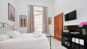 A hotel room with a bed, a TV, a console table with a vase of flowers, and a view of buildings through the window. - 4bnb - Cavour Square Apartment (Rome)