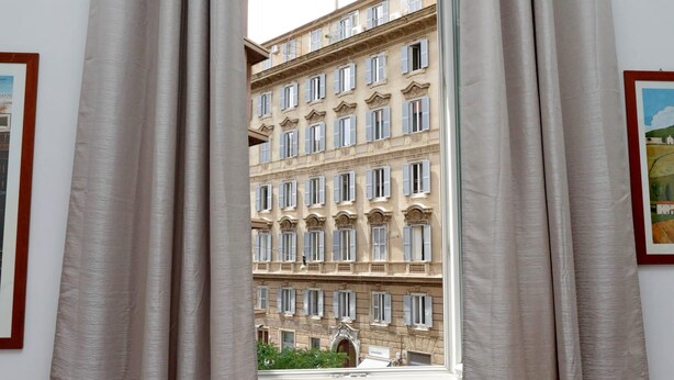 A view through a window with curtains, revealing a multi-story building with classical architecture and multiple windows. - 4bnb - Cavour Square Apartment (Rome)