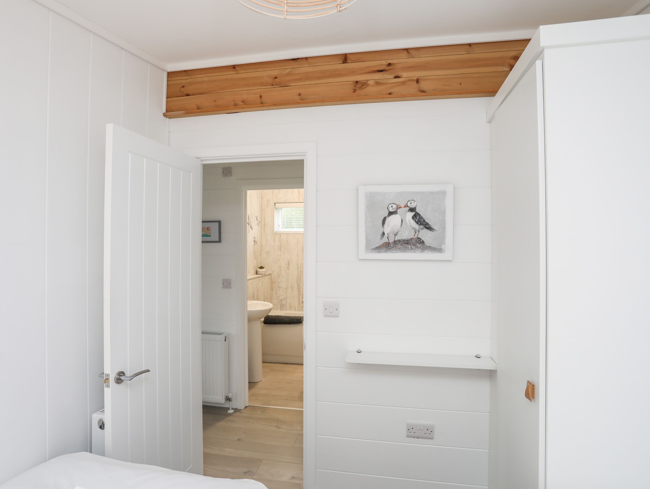 A narrow hallway with white walls and wooden beams, leading to a bathroom with a white sink and a framed picture of birds on the wall.