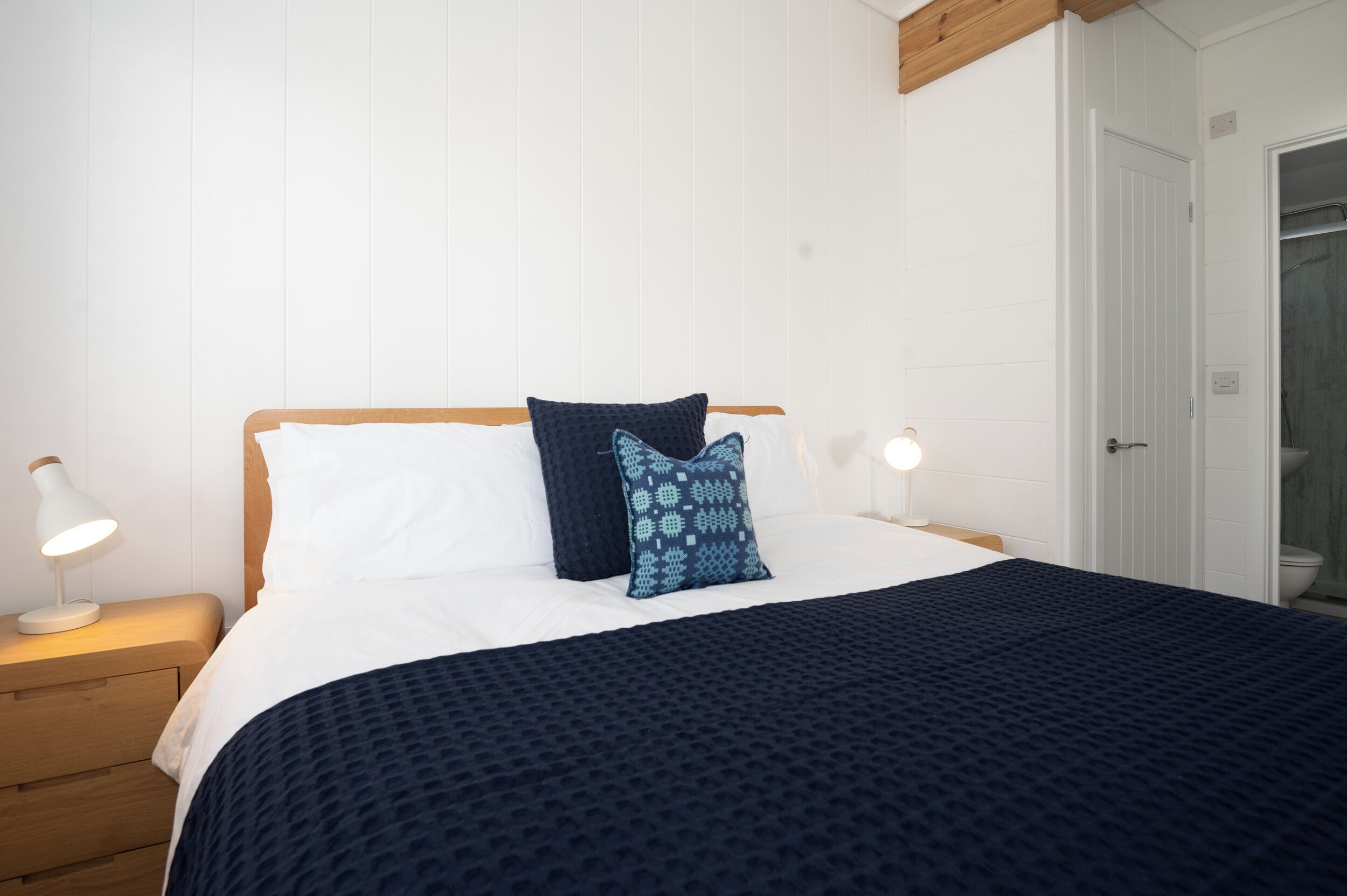 Een net opgemaakt bed met een donkerblauwe deken en kussens, een houten nachtkastje met een lamp, en een witte deur op de achtergrond.