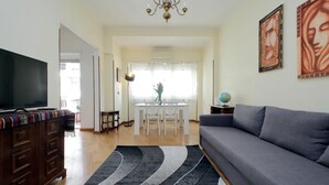 Uma sala de estar com sofá cinza, uma televisão sobre um armário de madeira, um candelabro e uma área de jantar com mesa e cadeiras.