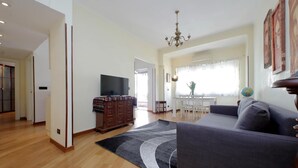 Uma sala de estar espaçosa com sofá cinza, piso de madeira, televisão de tela plana, canto de refeições com mesa e cadeiras, e um candelabro.