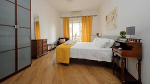 Quarto de hotel com cama, mesa de cabeceira, abajur e vista para o exterior através de uma janela com cortinas amarelas.