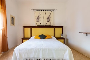 Uma cama bem arrumada com colcha branca e dois travesseiros, uma pequena mesa de cabeceira com abajur, e cortina fixada na parede com estampas de girafa.