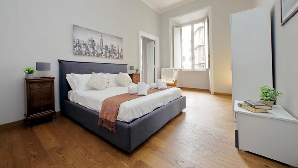 A modern bedroom with a large bed, a bedside table, a nightstand, a window, and a cityscape painting on the wall. - 4bnb - Spacious Belli Apartment (Rome)