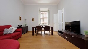 A living room with a red sofa, a dining table with chairs, a TV on a cabinet, and a window with a view of a building. - 4bnb - Spacious Belli Apartment (Rome)