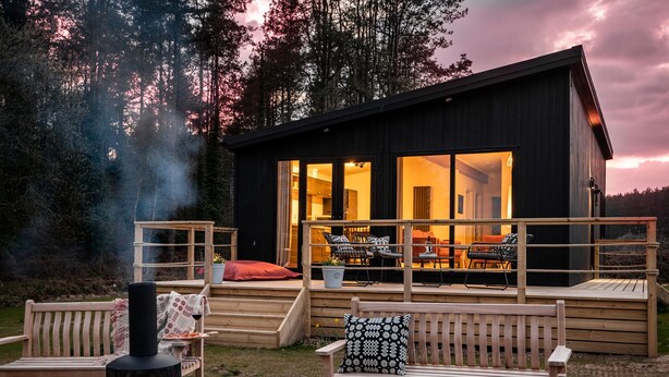 A cozy outdoor seating area with a fire pit, wooden benches, and a view of a modern cabin with large windows and a deck. - Murmur y Coed (Llanfairpwllgwyngyll)