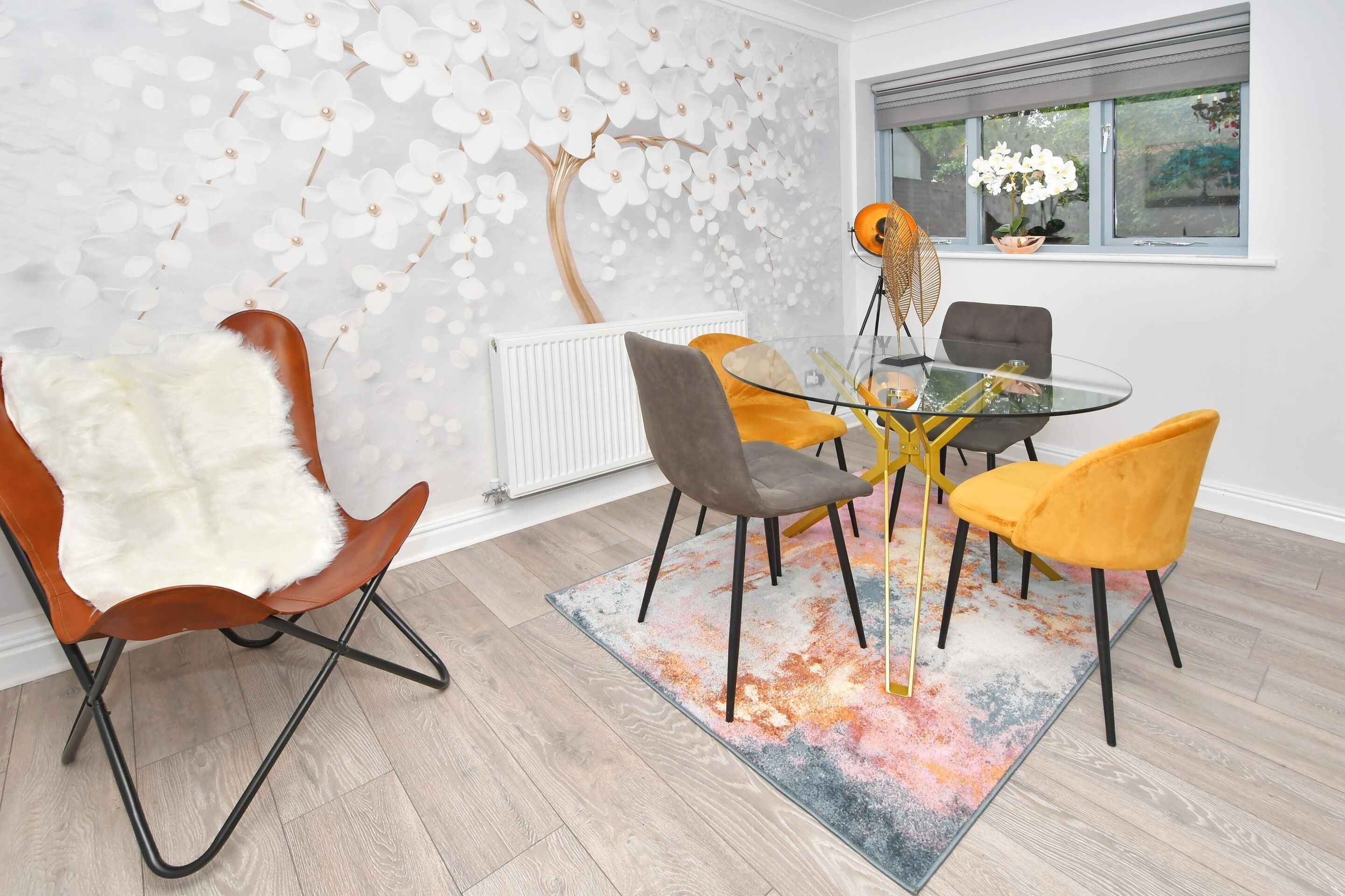 Un espace repas moderne avec une table en verre, des chaises orange et grises, un fauteuil blanc à poils longs et un mur décoratif orné d’un arbre et de fleurs.