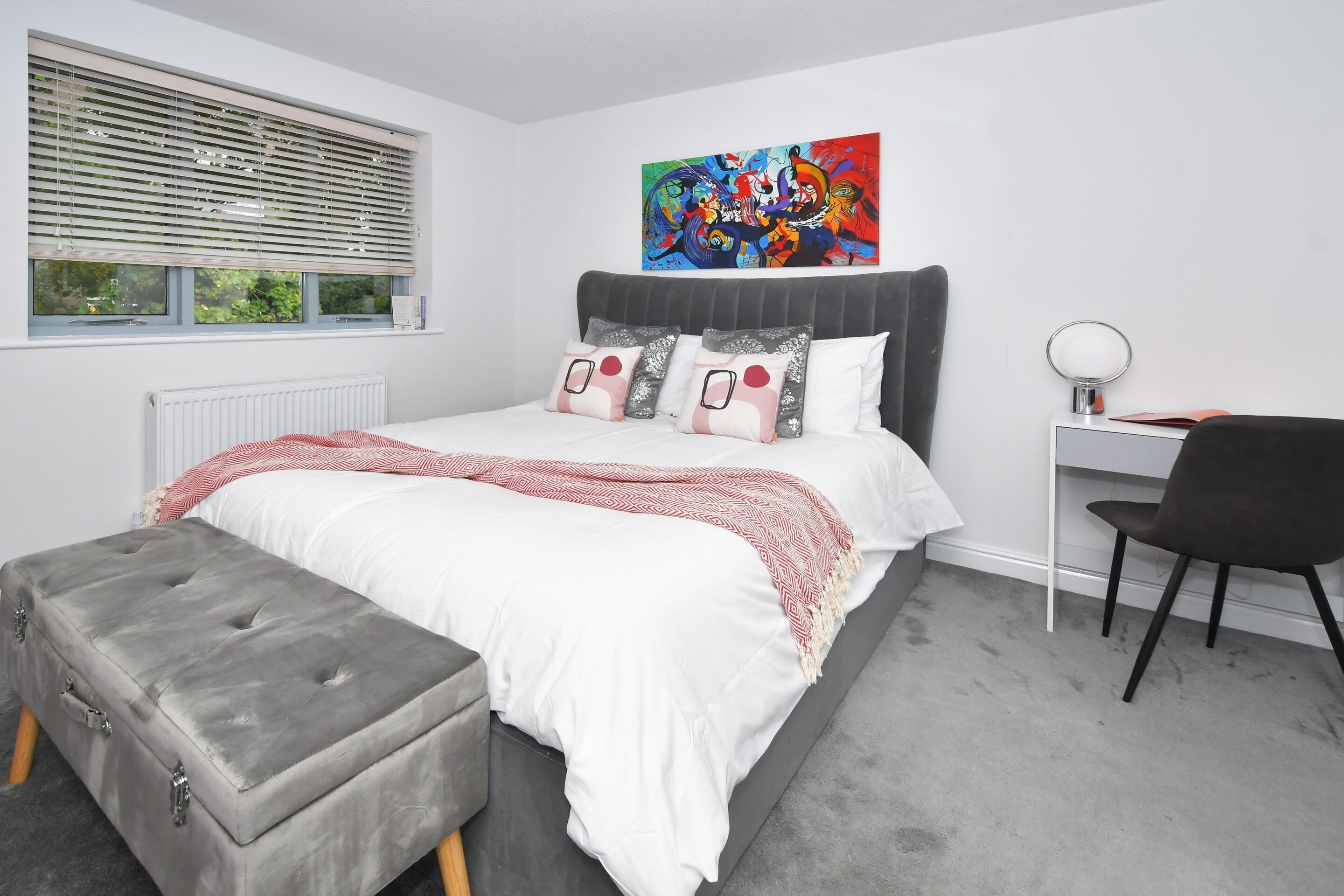 Een moderne slaapkamer met een grijs gestoffeerde bank, een grijs hoofdeinde en een kleurrijk abstract schilderij boven het bed.