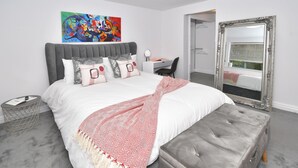 A modern bedroom with a large bed, a grey upholstered bench, a desk, and a mirror.