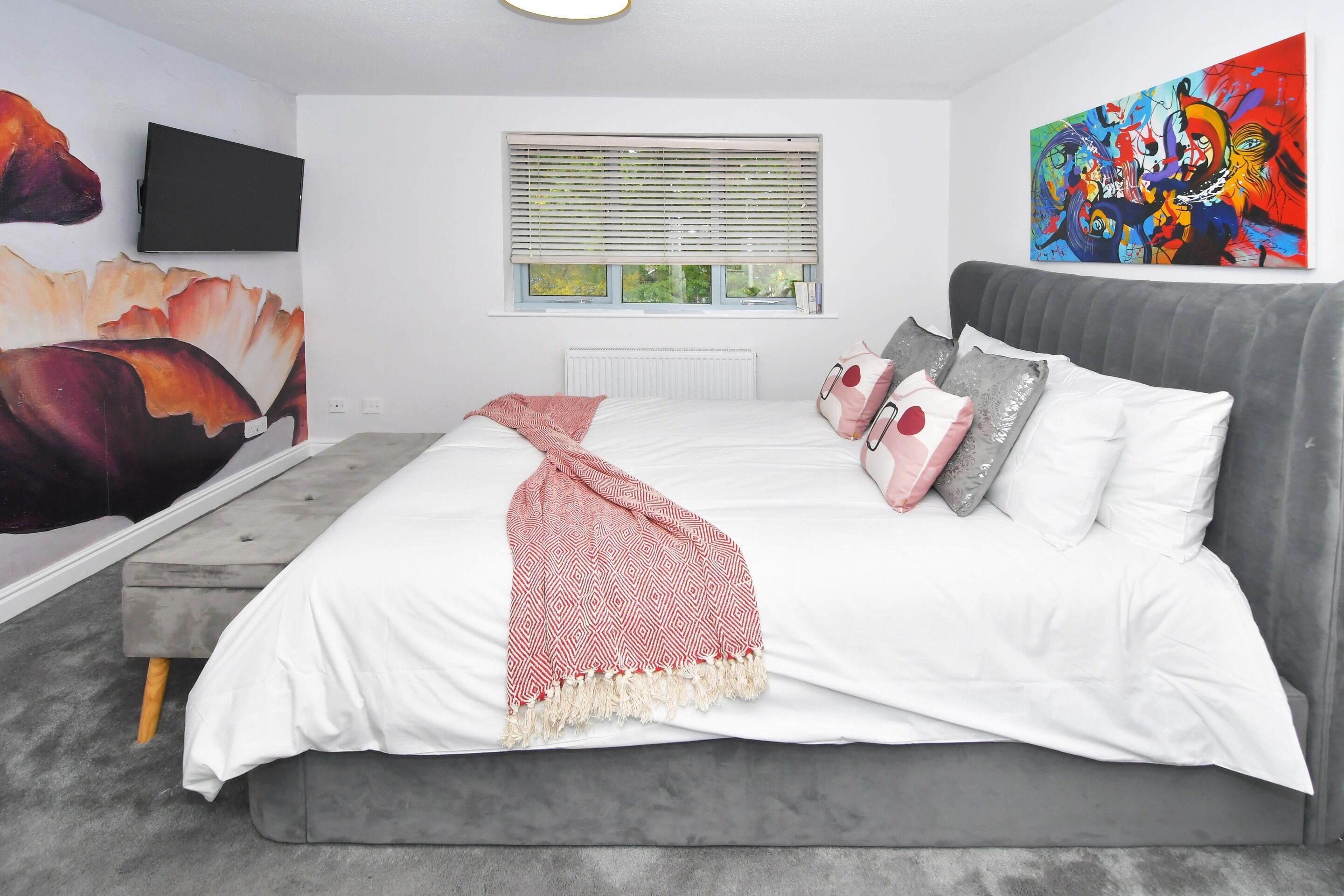 A bedroom with a bed, a grey sofa, a window with blinds, a painting on the wall, and a flat-screen TV mounted above the bed.