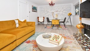 A living room with a sofa, a round coffee table with a book, a dining area with chairs, a chandelier, and a TV.