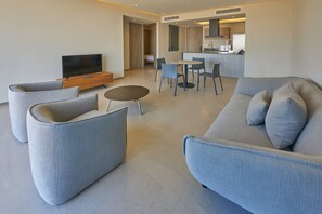Un salon moderne avec un canapé gris, deux fauteuils, une table basse et un téléviseur posé sur un meuble en bois. On aperçoit à l’arrière-plan un coin cuisine avec une table et des chaises.