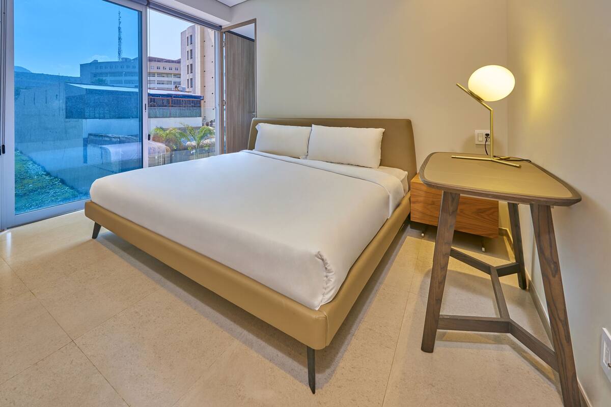 A modern hotel room with a large bed, a bedside table with a lamp, and a view of the city through a large window.