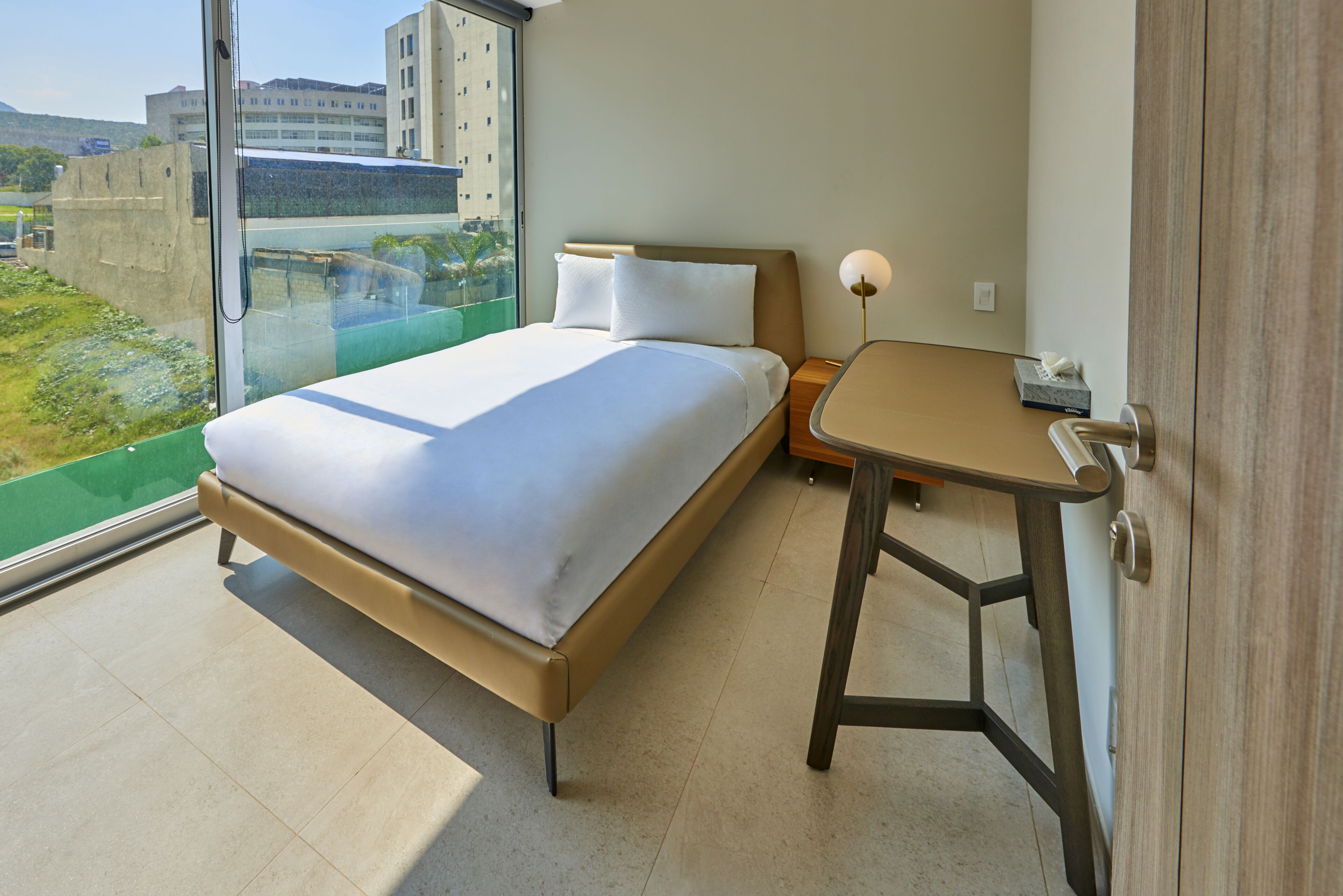 Une chambre d’hôtel moderne, dotée d’un grand lit, d’une petite table et offrant une vue sur le paysage urbain grâce à une grande fenêtre.