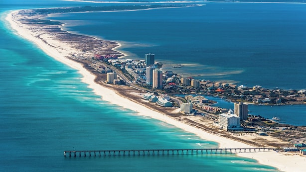 Vista aerea di una città costiera con una lunga spiaggia sabbiosa, un molo che si protende nell'acqua e diversi palazzi alti lungo la costa.