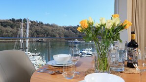 Ein Esstisch ist gedeckt mit weißen Tellern, Schüsseln und Gläsern, einer Weinflasche und einer Vase mit gelben Blumen. Ein Stuhl steht an der Tischplatte, und durch eine Glasbrüstung ist ein Blick auf eine Marina mit Booten und einer Brücke zu sehen.